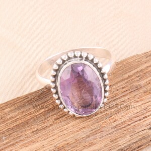 Pu&ograve; includere: Un anello d'argento con una pietra preziosa ovale di colore viola chiaro. La pietra &egrave; incastonata in una lunetta con un bordo di perline. L'anello &egrave; esposto su una superficie di legno.