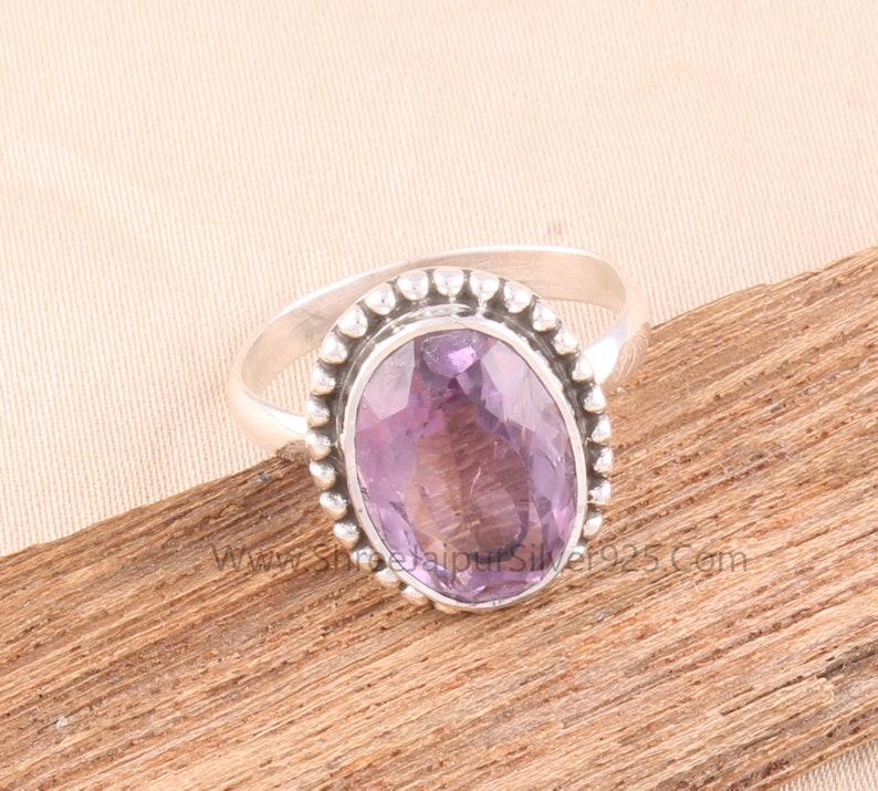 Pu&ograve; includere: Un anello d'argento con una pietra preziosa ovale di colore viola chiaro. La pietra &egrave; incastonata in una lunetta con un bordo di perline. L'anello &egrave; esposto su una superficie di legno.