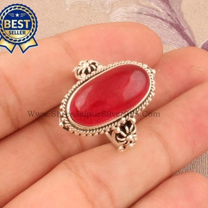 Puede incluir: Un anillo de plata con una piedra ovalada roja engastada en un diseño de filigrana decorativo. El anillo está en un dedo.