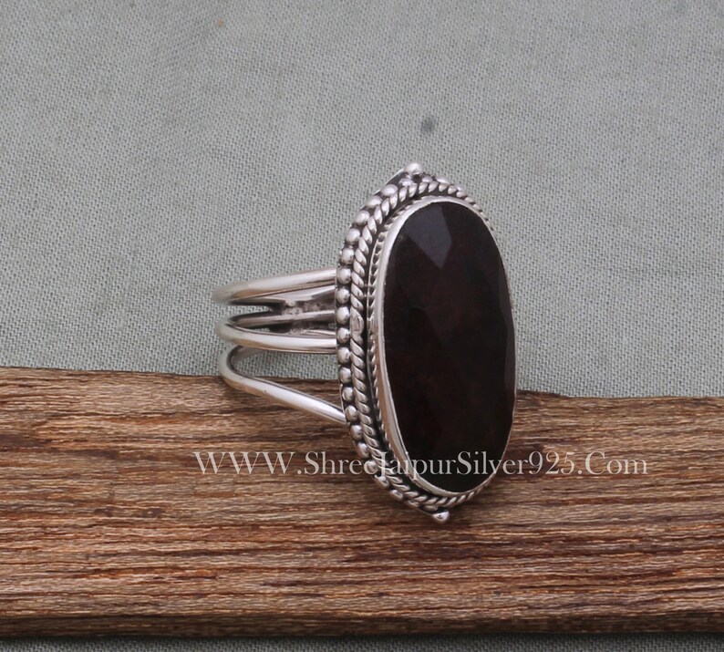 May include: A silver ring featuring a dark red oval gemstone set in a beaded bezel. The ring band has three bands.