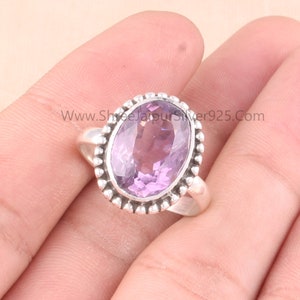 Pu&ograve; includere: Un anello in argento con una pietra preziosa ovale di colore viola chiaro. La pietra preziosa &egrave; incorniciata da un bordo di perline. L'anello &egrave; mostrato su una mano.