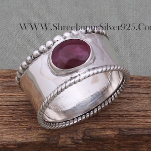 Op de afbeelding: Een zilveren ring met een rode edelsteen in een bezel-setting. De ring heeft een touwachtig ontwerp rond de band en een rij kralen rond de bovenkant.