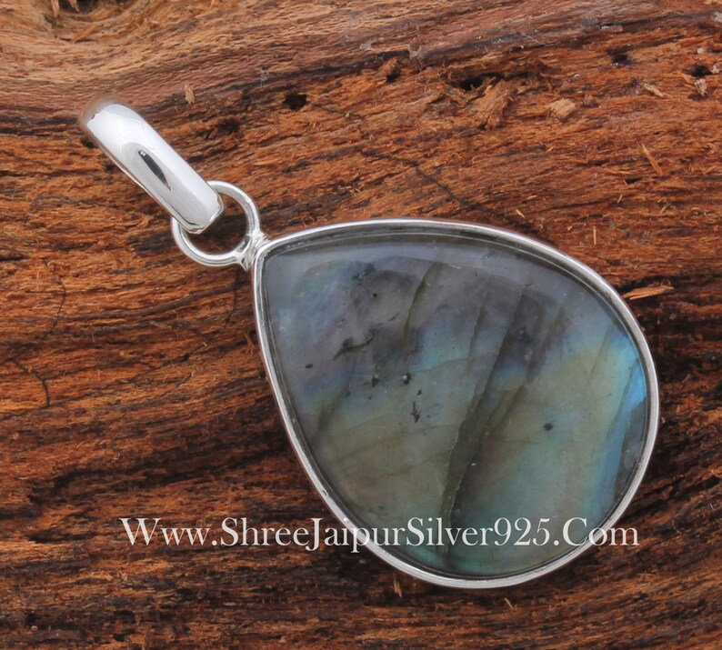 May include: A silver pendant with a teardrop-shaped labradorite gemstone. The gemstone has a blue-green iridescent sheen. The pendant is on a wooden surface.