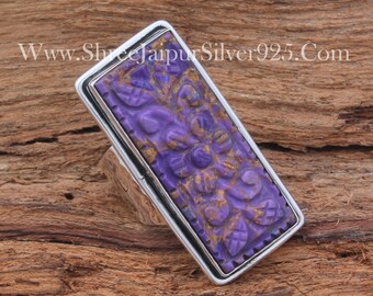 May include: A silver ring with a rectangular purple stone set in the center. The stone is carved with a floral design. The ring is on a wooden surface. www.shreejaipursilver925.com