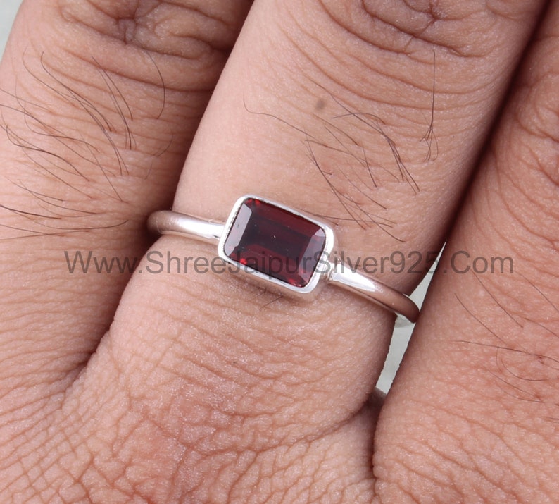 May include: A silver ring with a rectangular red gemstone. The ring is on a finger.