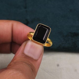May include: A gold-toned ring featuring a large, rectangular, faceted black gemstone set in a smooth metal bezel. The band is simple and circular, showcasing a polished finish that contrasts with the deep, dark, opaque surface of the stone.
