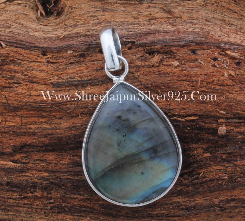 May include: A teardrop-shaped pendant with a silver setting and a blue and green labradorite gemstone. The pendant is on a wooden surface.