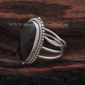 May include: A silver ring featuring a dark red oval gemstone set in an ornate bezel. The ring has a triple band design.