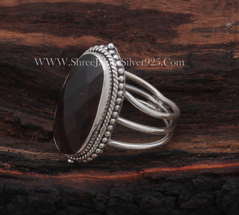 May include: A silver ring featuring a dark red oval gemstone set in an ornate bezel. The ring has a triple band design.