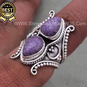 May include: A silver ring with two purple and white stones set in a detailed design. The ring is worn on a finger.
