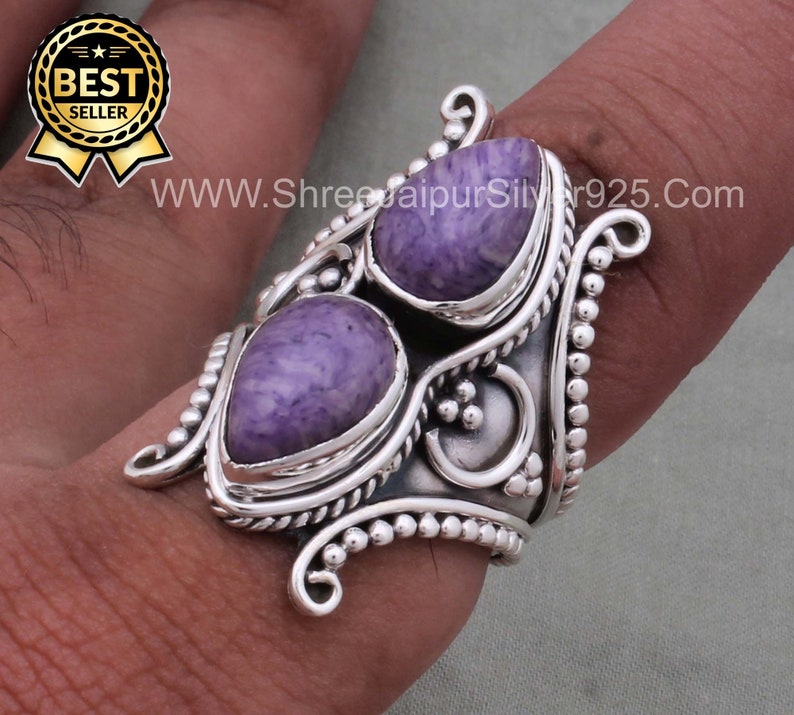 May include: A silver ring with two purple and white stones set in a detailed design. The ring is worn on a finger.
