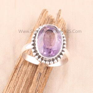 Pu&ograve; includere: Un anello d'argento con una grande ametista ovale. La gemma viola chiaro &egrave; incastonata in un bordo di perline. L'anello &egrave; esposto su un pezzo di legno. Il sito web www.ShreejaipurSilver925.com &egrave; visibile.