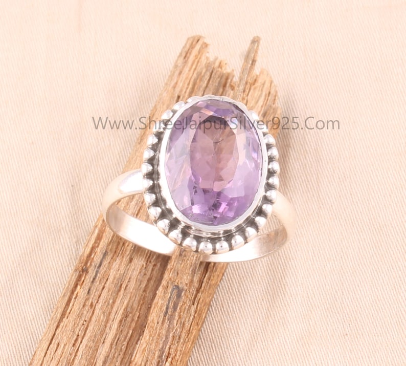 Pu&ograve; includere: Un anello d'argento con una grande ametista ovale. La gemma viola chiaro &egrave; incastonata in un bordo di perline. L'anello &egrave; esposto su un pezzo di legno. Il sito web www.ShreejaipurSilver925.com &egrave; visibile.