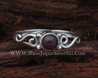 May include: A silver ring with a red gemstone set in a delicate, swirling design. The ring is on a wooden surface.