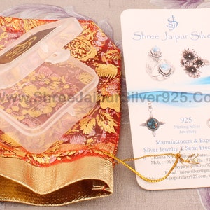 Pu&ograve; includere: Una scatola di gioielli in plastica trasparente aperta su un sacchetto a fantasia rosso e oro. &Egrave; visibile un biglietto da visita con immagini di gioielli e la scritta "Shree Jaipur Silver". Viene visualizzato anche l'indirizzo del sito web.