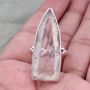 以下が含まれることがあります： 先のとがった形の透明な水晶の石がついた銀の指輪。石は、テクスチャ加工が施された銀のバンドにセットされています。指輪は手にしています。