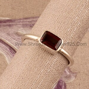 May include: A silver ring with a rectangular, dark red gemstone. The ring is on a brown fabric background. www.Shreesilver925.com