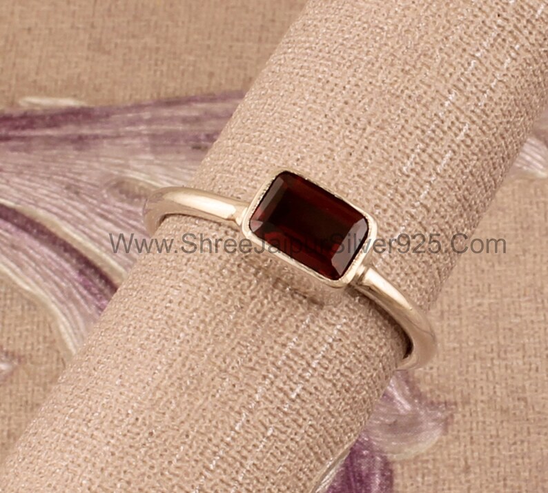 May include: A silver ring with a rectangular, dark red gemstone. The ring is on a brown fabric background. www.Shreesilver925.com