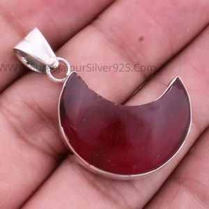 Pode incluir: Um pingente de prata com a forma de uma lua crescente com uma pedra preciosa vermelha escura. O pingente é feito de prata de lei e tem um acabamento polido.