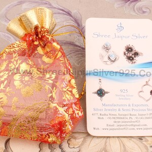 May include: A red and gold drawstring bag with a floral pattern. The bag is next to a white card with the text "Shree Jaipur Silver" and "925 Sterling Silver Jewellery" printed on it. The card also includes the company's contact information.