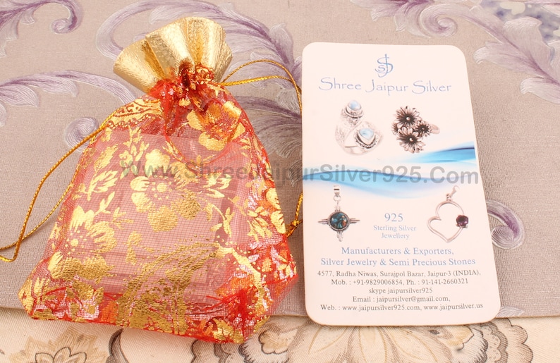 May include: A red and gold drawstring bag with a floral pattern. The bag is next to a white card with the text "Shree Jaipur Silver" and "925 Sterling Silver Jewellery" printed on it. The card also includes the company's contact information.
