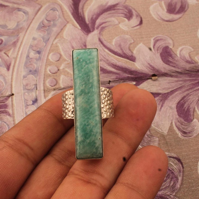 Op de afbeelding: Een rechthoekige, lichtgroene edelsteenring met een getextureerde zilveren band. De edelsteen is verticaal gezet in een zilveren frame. De ring wordt door een hand omhoog gehouden.