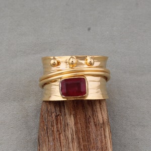 Pode incluir: Anel de ouro com uma gema vermelha em forma quadrada. O anel tem uma banda texturizada com três detalhes dourados.