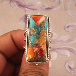 May include: A rectangular silver ring featuring a vibrant stone with turquoise, orange, red, and gold hues. The stone is set within a detailed silver frame, showcasing a unique and colorful gemstone.