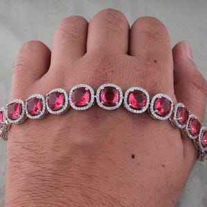Pode incluir: Uma pulseira de prata com grandes pedras preciosas vermelho rubi de corte quadrado. Cada rubi é rodeado por pequenos cristais transparentes. A pulseira é exibida numa mão.
