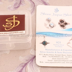May include: A white plastic box with a brown label that says "www.jaipursilver925.com" and a silver logo.  Next to the box is a white card with the text "Shree Jaipur Silver" and "www.ShreeJaipurSilver925.Com". The card also shows a silver pendant with a turquoise stone and a silver heart pendant with a purple stone.