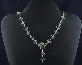 May include: A silver necklace with a green and red gemstone pendant. The necklace is made of small silver leaves with green gemstones set in them. The pendant is a flower with green gemstones and a red gemstone in the center.