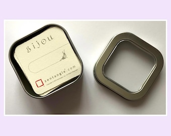 May include: A square metal tin with a clear window lid, containing a stack of off-white square cards. The top card features the text Bijou, a small snail illustration, and the text zentangle.com with a red square logo and copyright information.