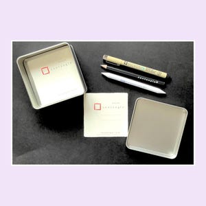 May include: A square metal tin with an open lid, a square paper tile with the red Zentangle logo, a beige Micron PN pen, a black Zentangle pencil, and a white paper blending stump. The paper tile features the text zentangle and zentangle.com.