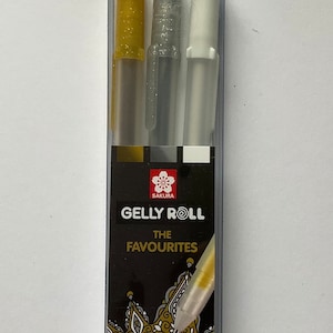 May include: A clear plastic package containing three gel pens. The pens have gold glitter, silver glitter, and white ink. The package has the text "Gelly Roll The Favourites" and "Fun Writing, Colouring and More!"