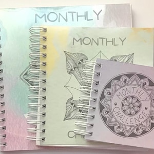 Könnte beinhalten: Ein Stapel spiralgebundener Notizbücher mit dem Wort "MONTHLY" auf den Umschlägen. Die Notizbücher haben Aquarellhintergründe in Pastellfarben und dekorative Strichzeichnungen. Ein kleineres Notizbuch zeigt den Text "MONTHLY CHALLENGE".