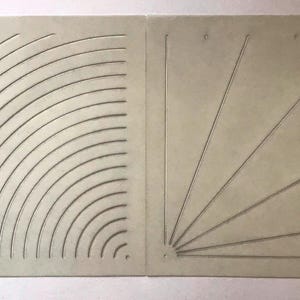 Könnte beinhalten: Zwei quadratische, hellbeige Platten mit eingravierten Mustern. Die linke Platte zeigt eine Reihe konzentrischer Viertelkreisbögen. Die rechte Platte weist eine Reihe gerader Linien auf, die von einem Punkt unten links ausgehen.