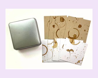 May include: A square silver metal tin container positioned next to a collection of square cards. The cards feature a mix of light brown and white backgrounds, each decorated with brown circular coffee ring stains and scattered brown paint splatters.