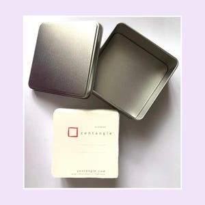 May include: A square silver metal tin with a separate lid and an open base. Beside the tin is a stack of off-white square paper tiles. The top tile features a red square logo, the text "an original zentangle", two lines, and "zentangle.com".