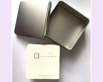 May include: A square silver metal tin with a separate lid and an open base. Beside the tin is a stack of off-white square paper tiles. The top tile features a red square logo, the text "an original zentangle", two lines, and "zentangle.com".