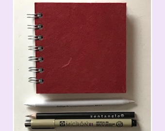 May include: A square, red, spiral-bound sketchbook with a textured cover. Below the sketchbook are three drawing tools: a white paper blending stump, a black Zentangle pencil, and a tan Pigma Micron 01 archival ink pen with black text.