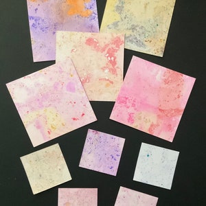 May include: Nine square pieces of paper with different watercolour patterns in shades of pink, purple, yellow, and orange. The paper is textured and has a speckled appearance.