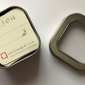 Könnte beinhalten: Eine silberne Metalldose mit einem durchsichtigen Plastikfenster auf dem Deckel. Die Dose enthält einen Stapel kleiner weißer Karten mit dem Wort "Bijou" und dem Text "zentangle.com" darauf.
