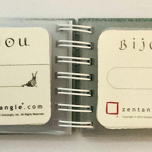 May include: A small, spiral-bound notebook with a green cover. The front page has the word "Bijou" written in black cursive. The page also has a small drawing of a snail and the text "zentangle.com".