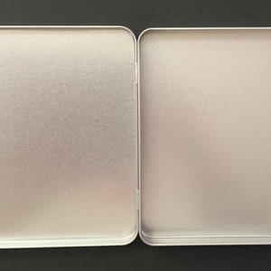 May include: An open, rectangular, silver-colored tin with rounded corners. The tin is divided into two equal halves, revealing a plain, textured interior. The metal has a matte finish, reflecting ambient light.