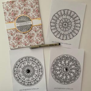 May include: A collection of mandala art cards and a floral notebook. The cards display detailed black and white circular patterns with the website address www.tangledwebcreations.com. A black pen rests on the cards. The notebook has a floral design and the text "Tangledweb Creations".