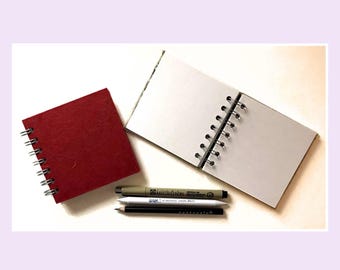 May include: A square notebook with a textured dark red cover and a silver wire spiral binding, alongside an open notebook with blank white pages, a tan Micron 01 pen, a white paper blending stump, and a black Zentangle pencil.
