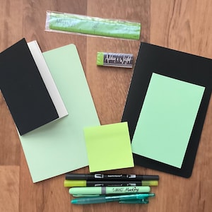 May include: An assortment of stationery items including a black notebook, green notepads, sticky notes, pens, and a ruler. The pens include BIC and Dual Art Marker brands. The text "Address & Wright" is visible on a small green item.