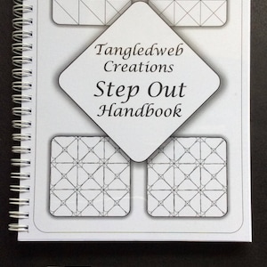 May include: A white spiral bound book with the title "Tangledweb Creations Step Out Handbook". The book has four black and white geometric patterns on the cover. Two black pens and a white pencil are lying on a black surface.