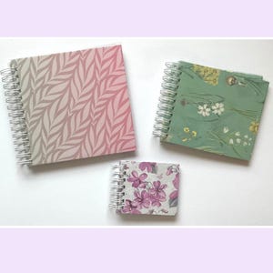 May include: Three spiral-bound notebooks of varying sizes. The largest features a pink and beige leaf pattern. The medium notebook is green with a floral print. The smallest notebook displays a light gray background with purple flower illustrations.