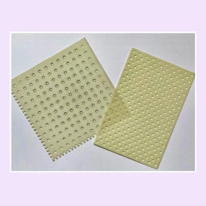Könnte beinhalten: Zwei rechteckige Platten aus blassgelbem Material mit einem gleichmäßigen Raster aus kleinen, kreisförmigen Perforationen. Eine Platte weist an einer Seite einen gezackten, kammartigen Rand auf, während die andere Platte glatte Kanten hat.